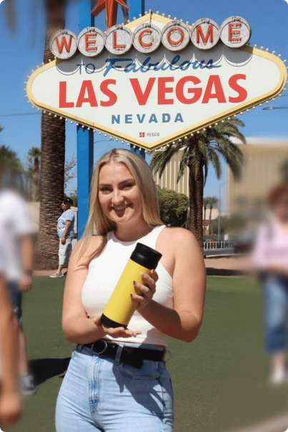 A woman holding the comfort sip prototype model in a yellow color and standing close to the sign post which says, 'Welcome to fabulous Las Vegas Nevada'
